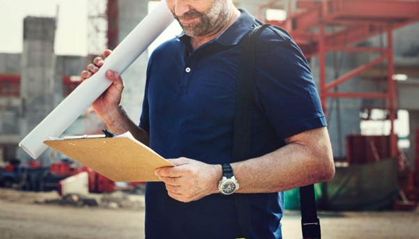 Commercial general contractor in a dark blue shirt holding a clipboard at a construction site.